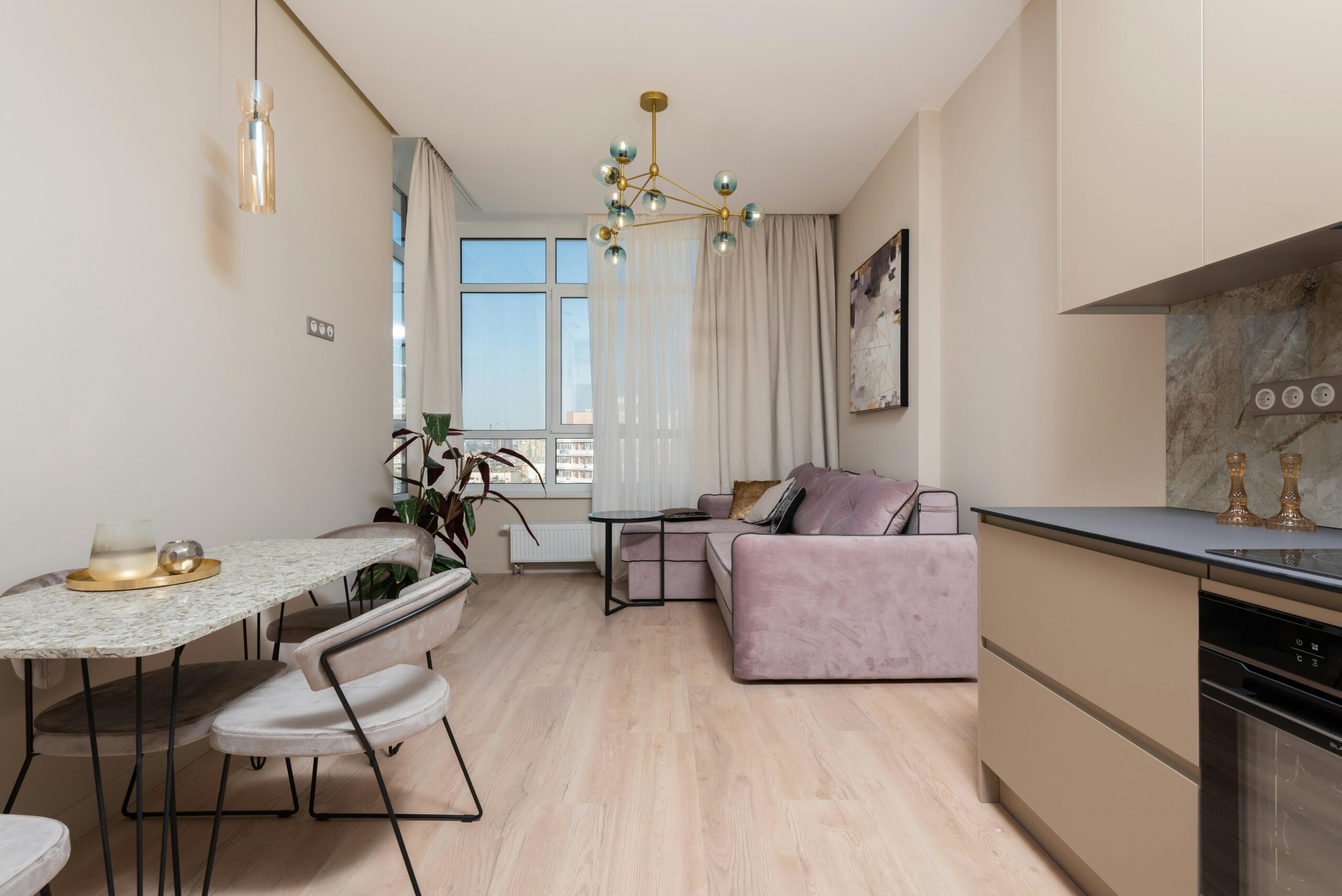 Interior of bright modern studio with open kitchen with cabinets near table and chairs next to couch with pillows near window with curtains